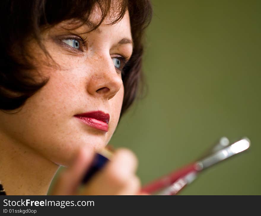 Brown-haired girl makes a make-up. Looks aside. Green background. Lipstick red. Eyes blue. Holds pencils. Low depth of field. Brown-haired girl makes a make-up. Looks aside. Green background. Lipstick red. Eyes blue. Holds pencils. Low depth of field.