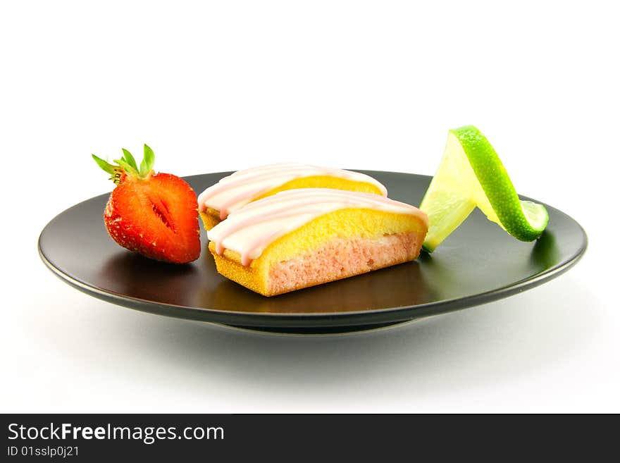 Two pink strawberry sliced cakes with icing on the top with a half strawberry and twist of lime on a black plate on a white background. Two pink strawberry sliced cakes with icing on the top with a half strawberry and twist of lime on a black plate on a white background