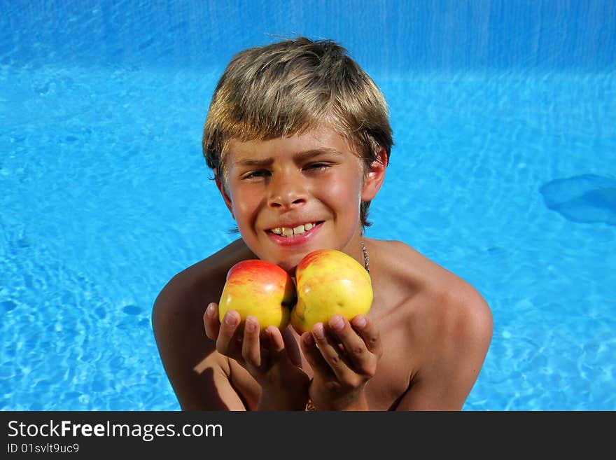 A 10 year old cute and smiling American - German boy sitting at a swimming pool and presenting delicious apples in the summer sun. A 10 year old cute and smiling American - German boy sitting at a swimming pool and presenting delicious apples in the summer sun