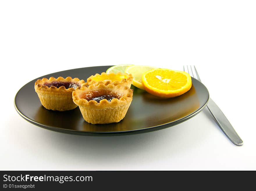 Red and yellow small jam tarts with slices of lemon, lime, and orange on a black plate with a fork on a white background. Red and yellow small jam tarts with slices of lemon, lime, and orange on a black plate with a fork on a white background