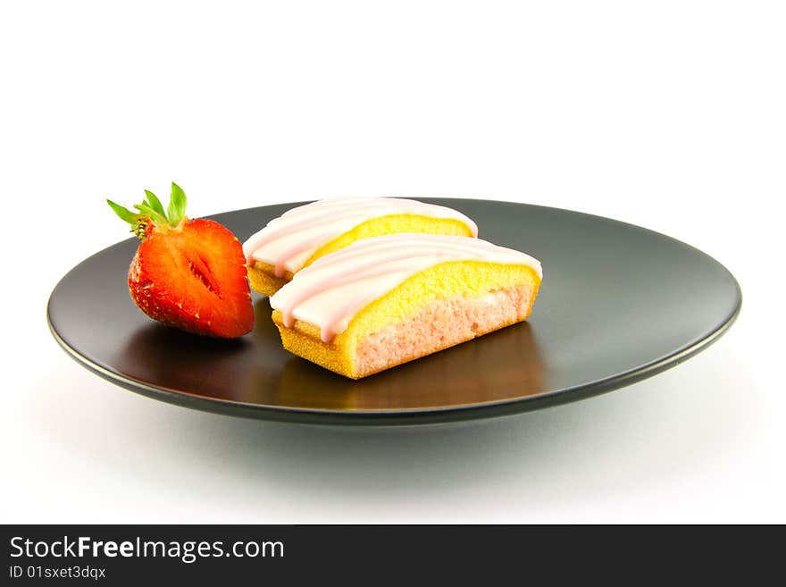 Two pink strawberry sliced cakes with icing on the top with a half strawberry on a black plate on a white background. Two pink strawberry sliced cakes with icing on the top with a half strawberry on a black plate on a white background