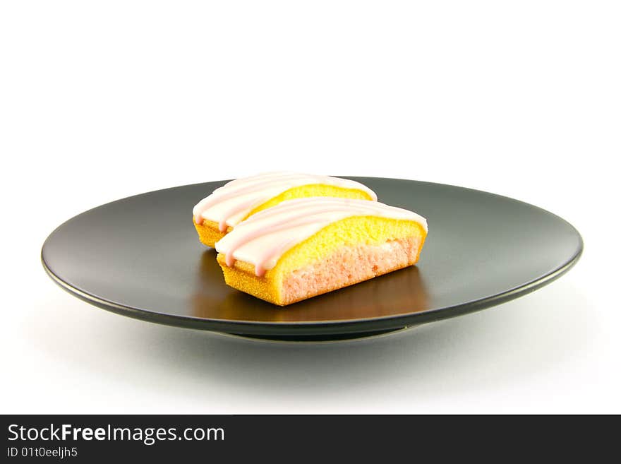 Two strawberry sliced cakes with icing on the top and pink and yellow sponge on a black plate with a white background. Two strawberry sliced cakes with icing on the top and pink and yellow sponge on a black plate with a white background