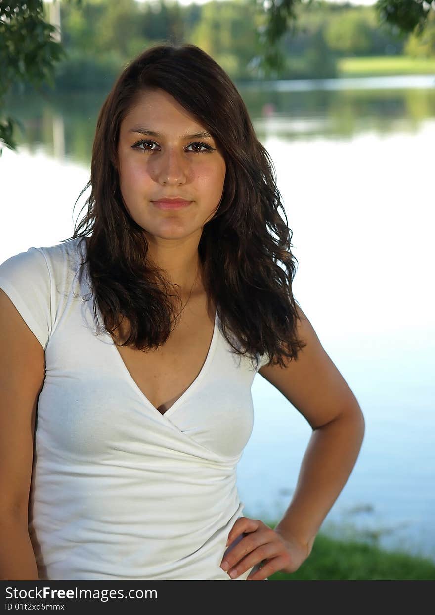 A beautiful young brunette woman standing by a serene lake with her hand on her hips. A beautiful young brunette woman standing by a serene lake with her hand on her hips.