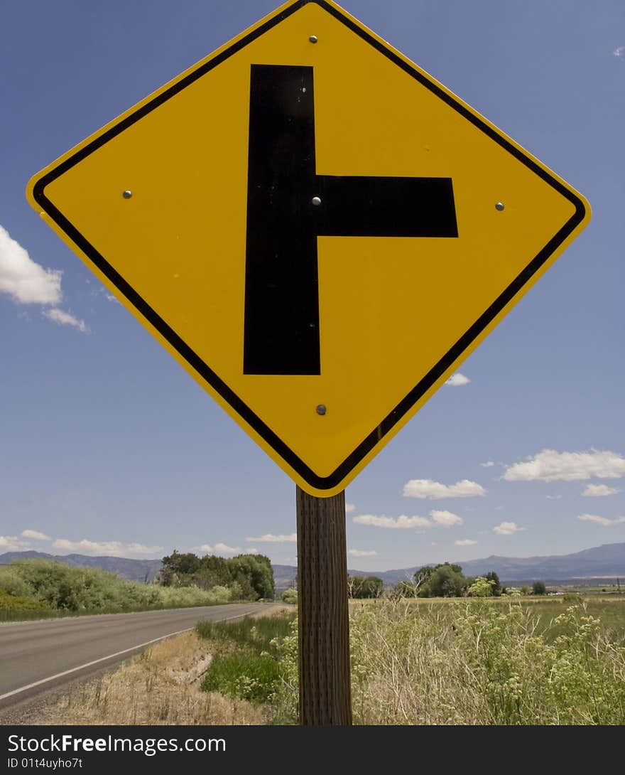 This is a road sign with a highway, mountains and a bright blue sky in the backgraound.