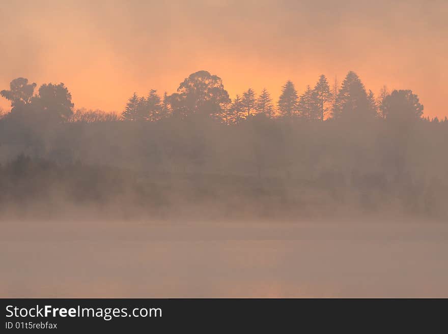 Early morning at a lake/dam with an orange glow visible through the trees and mist. Early morning at a lake/dam with an orange glow visible through the trees and mist