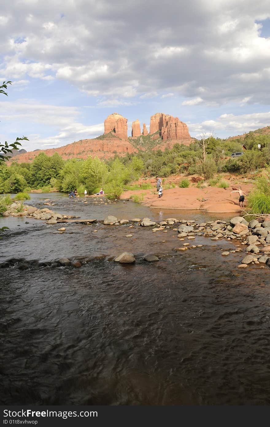 View of red rock formation, cathedral rock in sedona arizona, with small river running in the foregraound.  various people hiking and playing along riverbank