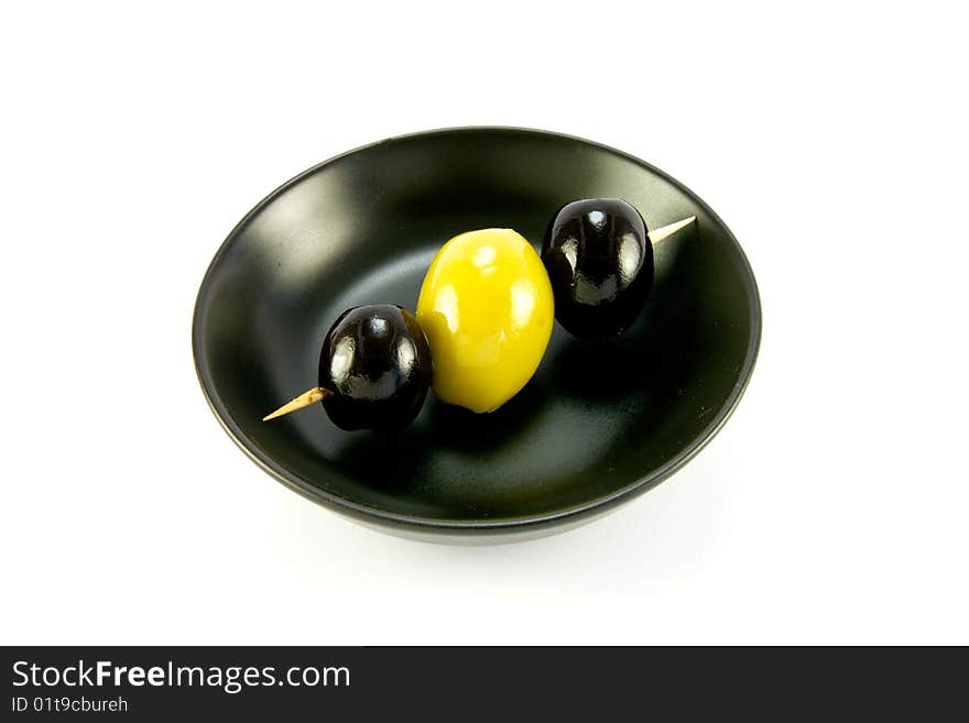 Two black and one green olive on a stick in a small black bowl on a white background. Two black and one green olive on a stick in a small black bowl on a white background