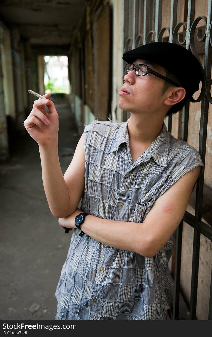 Bespectacled asian man in beret smoking while waiting along a corridor in a deserted abandoned place. Bespectacled asian man in beret smoking while waiting along a corridor in a deserted abandoned place