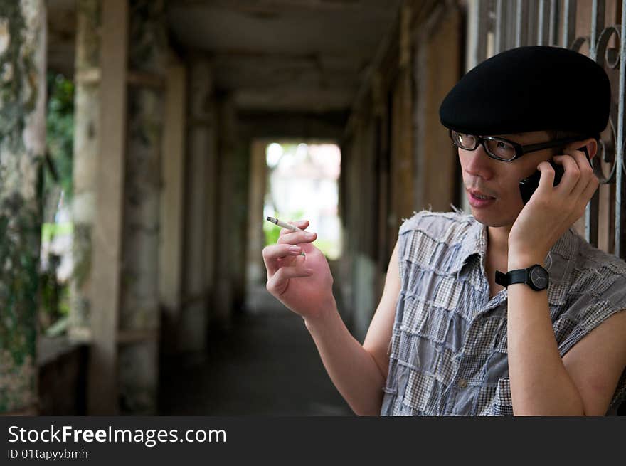 Bespectacled asian man in beret smoking and talking on the mobile phone along a corridor in a deserted abandoned place. Bespectacled asian man in beret smoking and talking on the mobile phone along a corridor in a deserted abandoned place