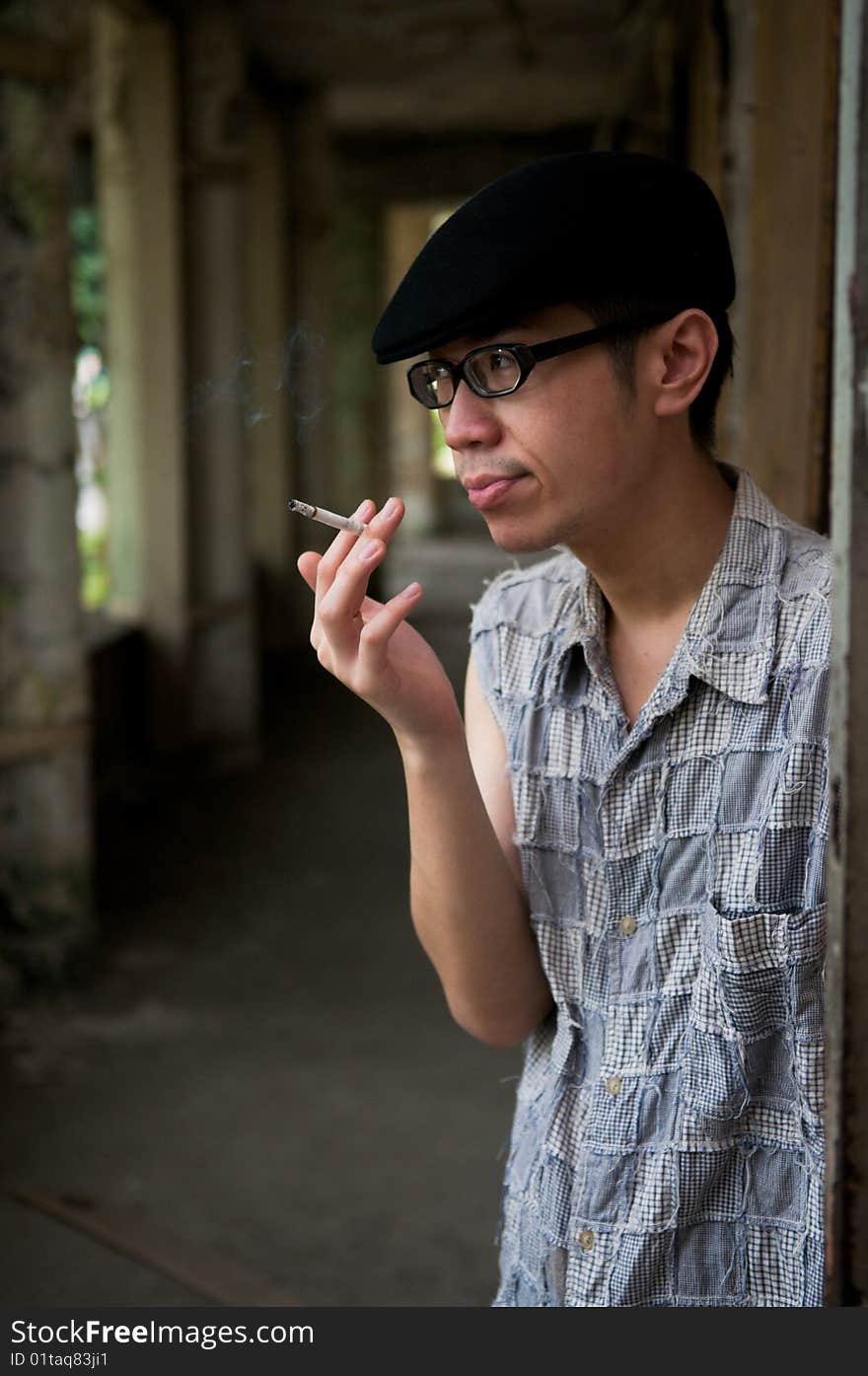 Frowning bespectacled asian man in beret smoking while waiting along a corridor in a deserted abandoned place