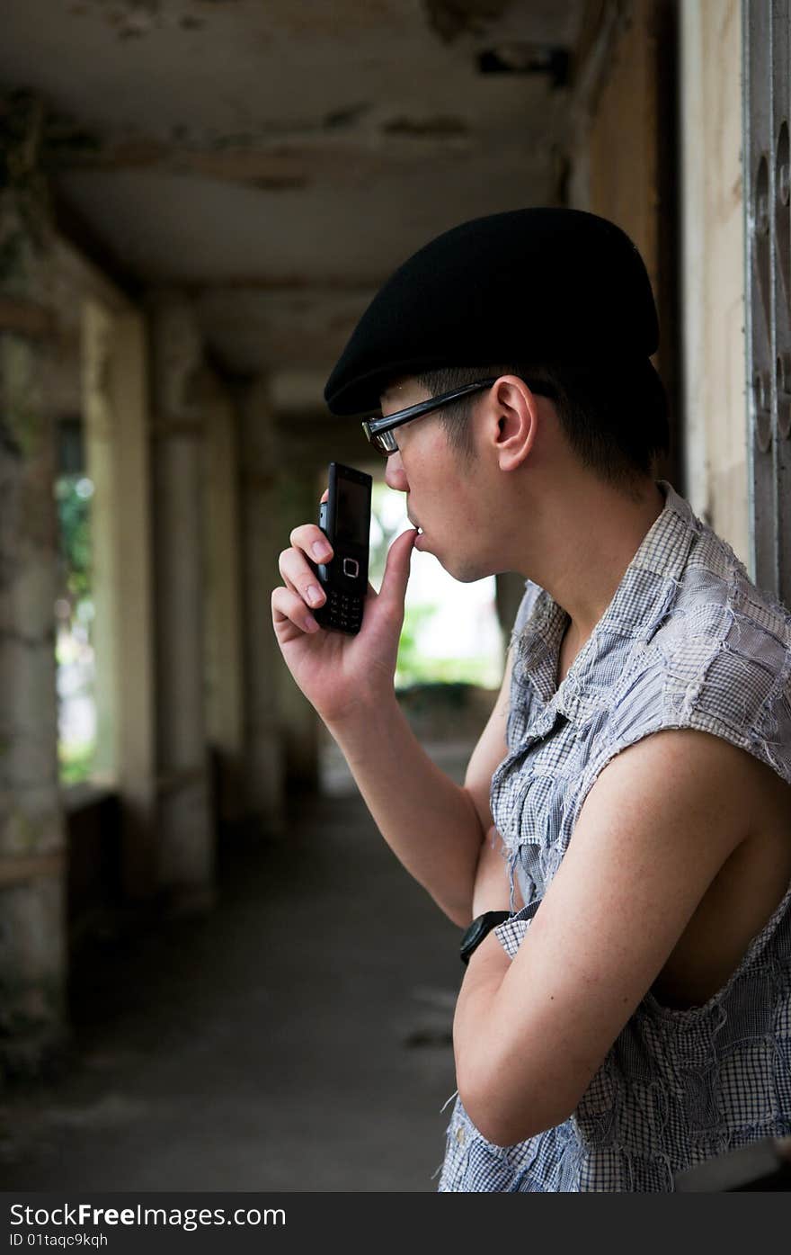 Bespectacled asian man in beret holding a mobile phone and with thumb on lips along a corridor in a deserted abandoned place. Bespectacled asian man in beret holding a mobile phone and with thumb on lips along a corridor in a deserted abandoned place