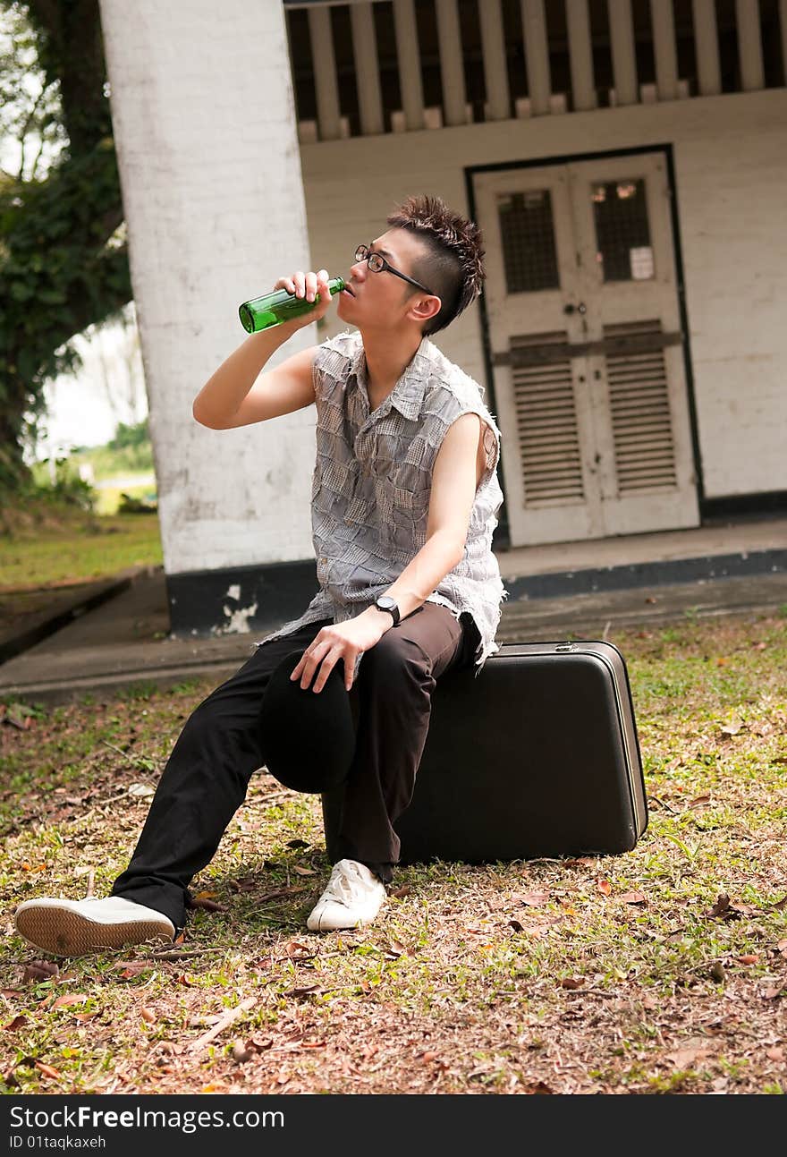 Bespectacled asian man drinking from a bottle and sitting on a suitcase outside the boarded up door of an abandoned apartment. Bespectacled asian man drinking from a bottle and sitting on a suitcase outside the boarded up door of an abandoned apartment