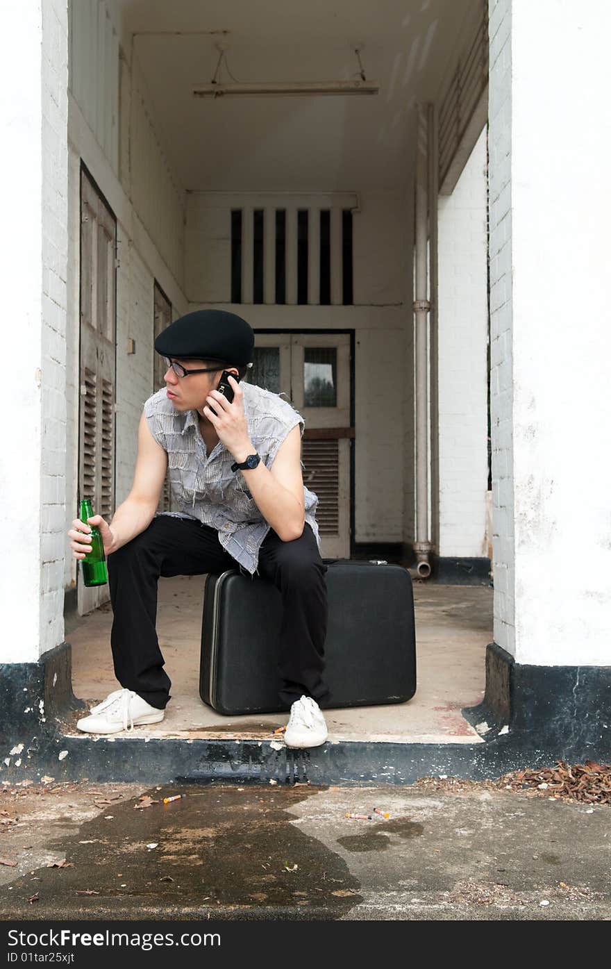 Bespectacled Asian man listening to the mobile phone while sitting on a suitcase at the end of a corridor of a boarded up and abandoned apartment. Bespectacled Asian man listening to the mobile phone while sitting on a suitcase at the end of a corridor of a boarded up and abandoned apartment