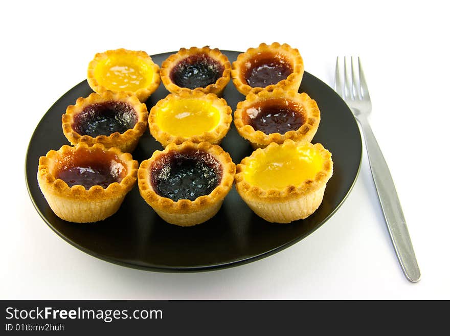 Selection red and yellow small jam tarts on a black plate with a fork on a white background