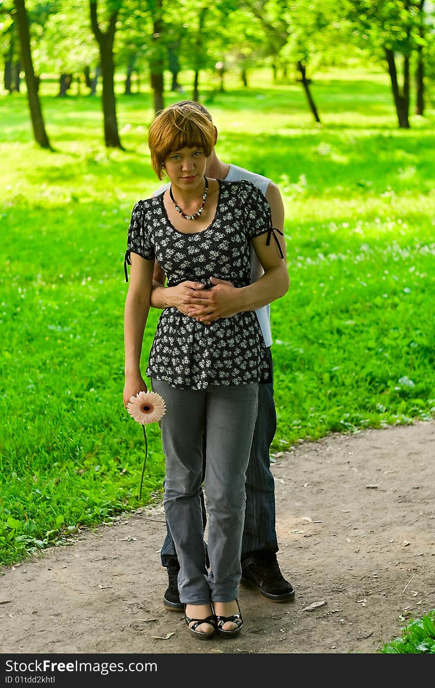 Teenagers: Man stands behind woman with flower and embraces she. It is on nature. Full-length portrait