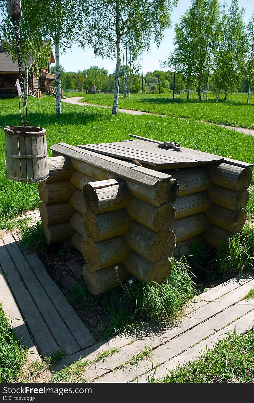 Wooden draw well in the rural area with shadoof and bucket. Green grass around it. Wooden house in background. Wooden draw well in the rural area with shadoof and bucket. Green grass around it. Wooden house in background