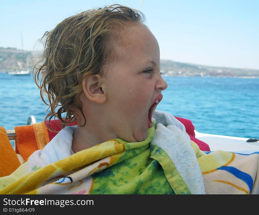 A young toddler sitting on a boat in the middle of the sea wrapped in a towel and yawning. A young toddler sitting on a boat in the middle of the sea wrapped in a towel and yawning