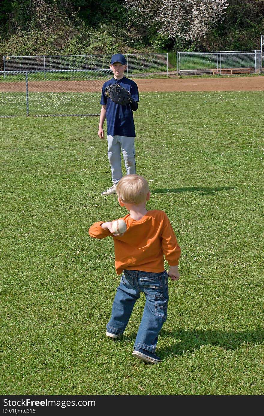 A 3 year old boy wearing orange shirt and blue jeans is throwing a baseball to his big brother between little league game. A 3 year old boy wearing orange shirt and blue jeans is throwing a baseball to his big brother between little league game.