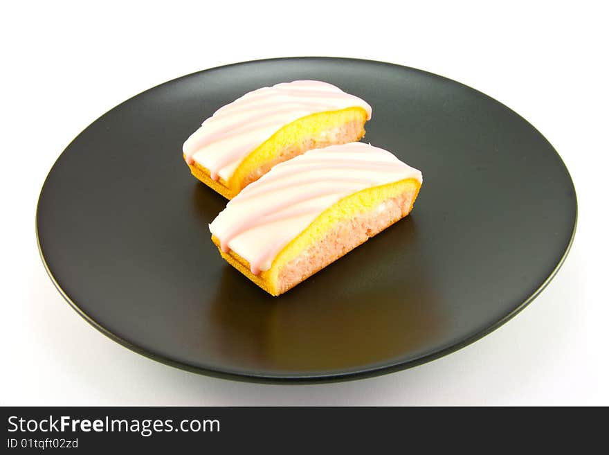 Two strawberry sliced cakes with icing on the top and pink and yellow sponge on a black plate with a white background. Two strawberry sliced cakes with icing on the top and pink and yellow sponge on a black plate with a white background