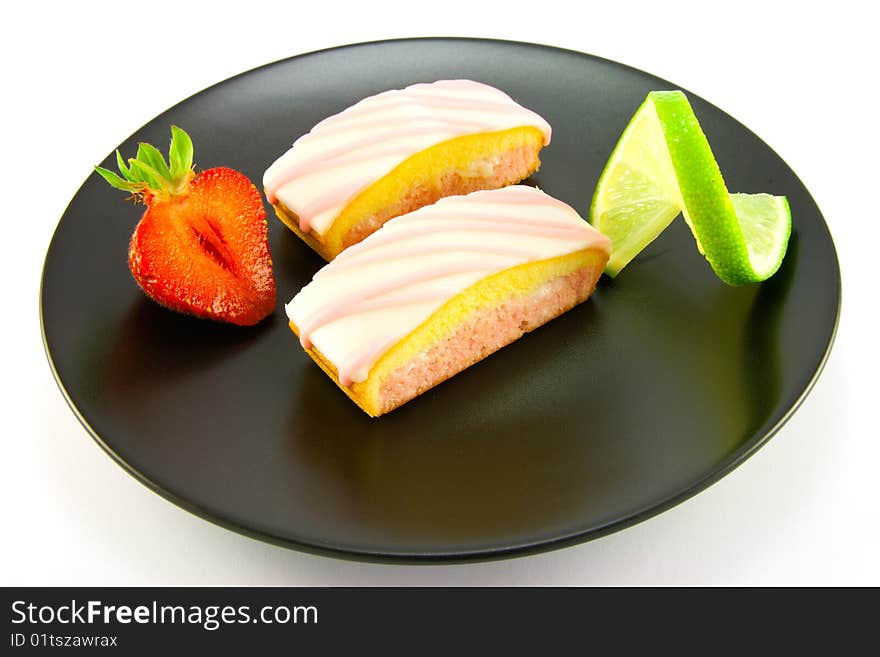 Two pink strawberry sliced cakes with icing on the top with a half strawberry and twist of lime on a black plate on a white background. Two pink strawberry sliced cakes with icing on the top with a half strawberry and twist of lime on a black plate on a white background