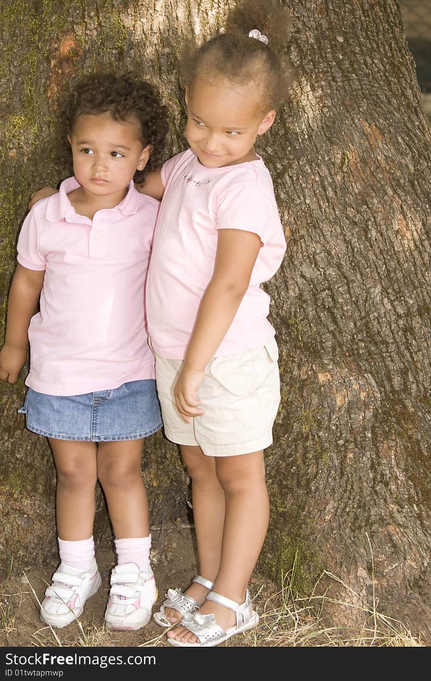 Two Young Ethnic Sisters with Questioning and Mischievous looks on their faces. They have their arms around each other as hold each other while standing against the trunk of a large tree.