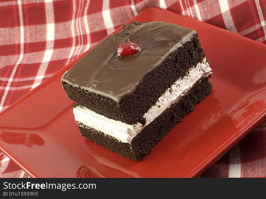 A piece of delicious chocolate layer cake with white frosting in the center layer, and chocolate frosting on top with a cherry, red plate with red plaid tablecloth