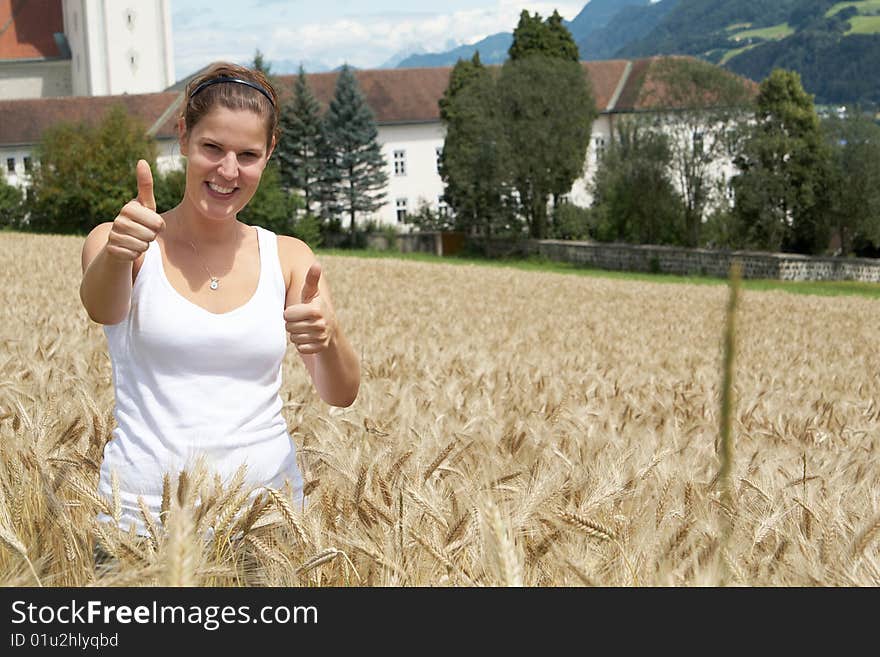 A young woman gives a thumbs up sign in the yellow field under blue sky in a rural alpine setting. Village in the background. Focus is on the woman. A young woman gives a thumbs up sign in the yellow field under blue sky in a rural alpine setting. Village in the background. Focus is on the woman.