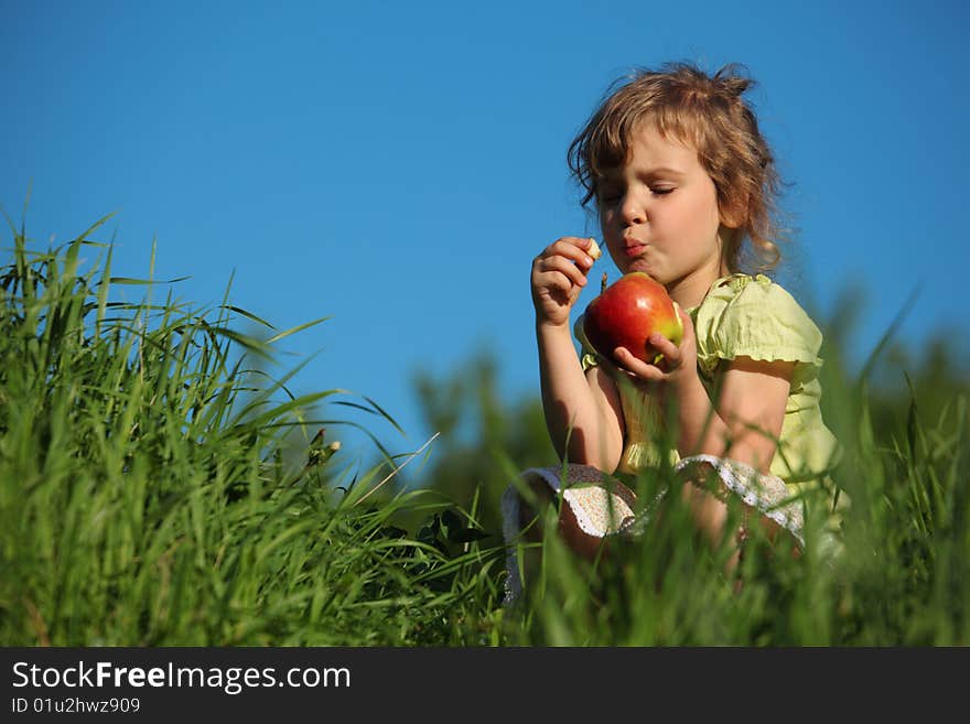 Girl eats red apple in grass against blue sky