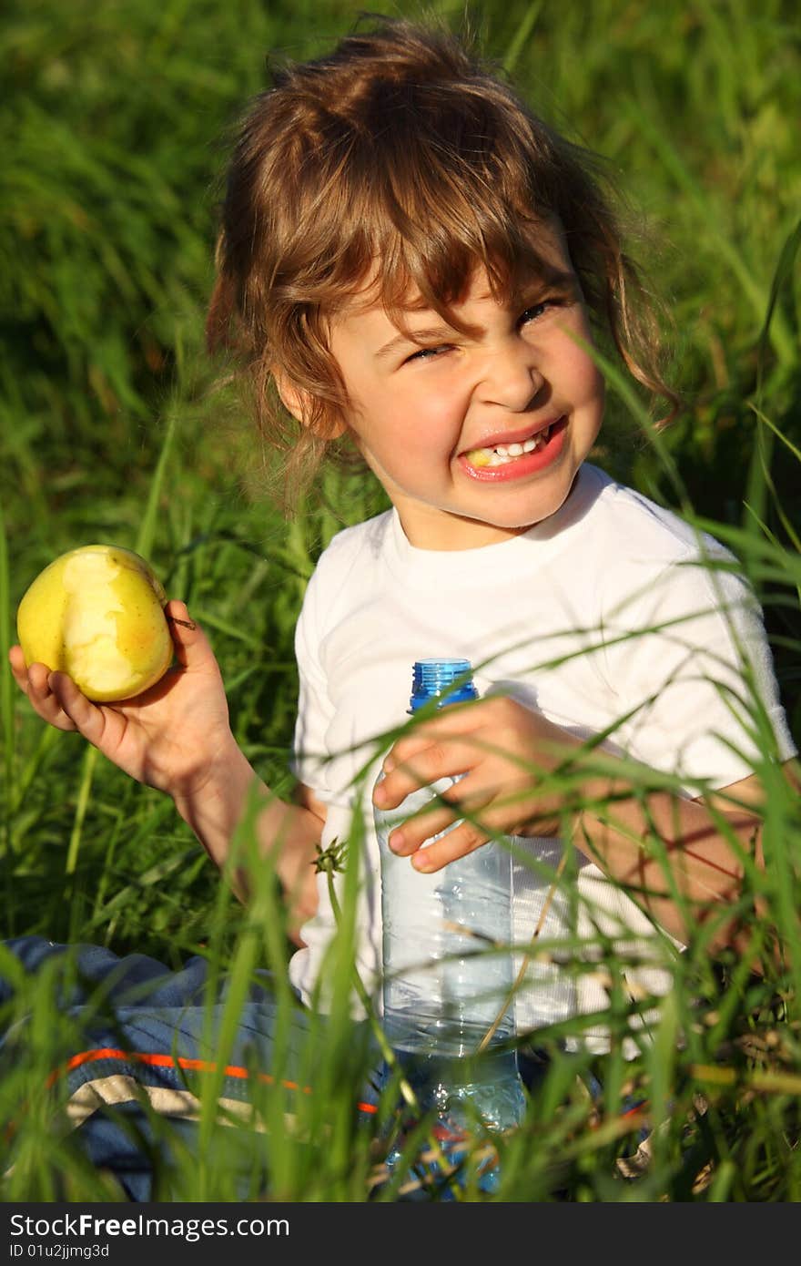 Girl with plastic bottle eats green apple in grass