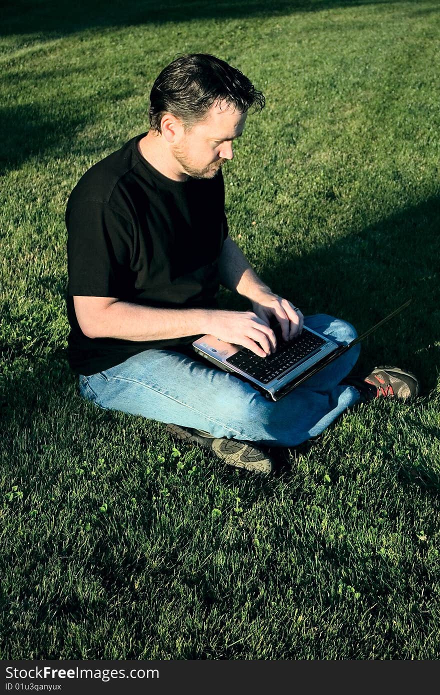 A young Caucasian man works with his laptop computer on the lush green grass. A young Caucasian man works with his laptop computer on the lush green grass.