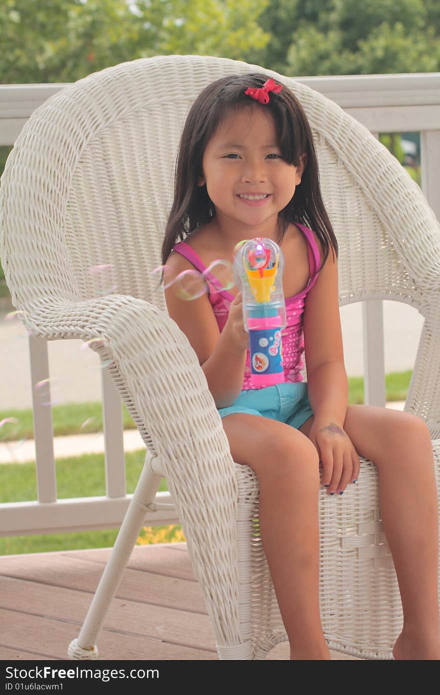 A pretty little Asian girl sits in a wicker chair, making bubbles with a bubble gun. A pretty little Asian girl sits in a wicker chair, making bubbles with a bubble gun.