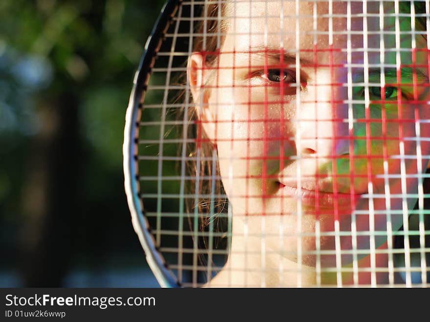 Young beautiful face through the badminton racket; lateral warm natural lighting; there is a colour sunbeam on the cheek in the shadow. Young beautiful face through the badminton racket; lateral warm natural lighting; there is a colour sunbeam on the cheek in the shadow