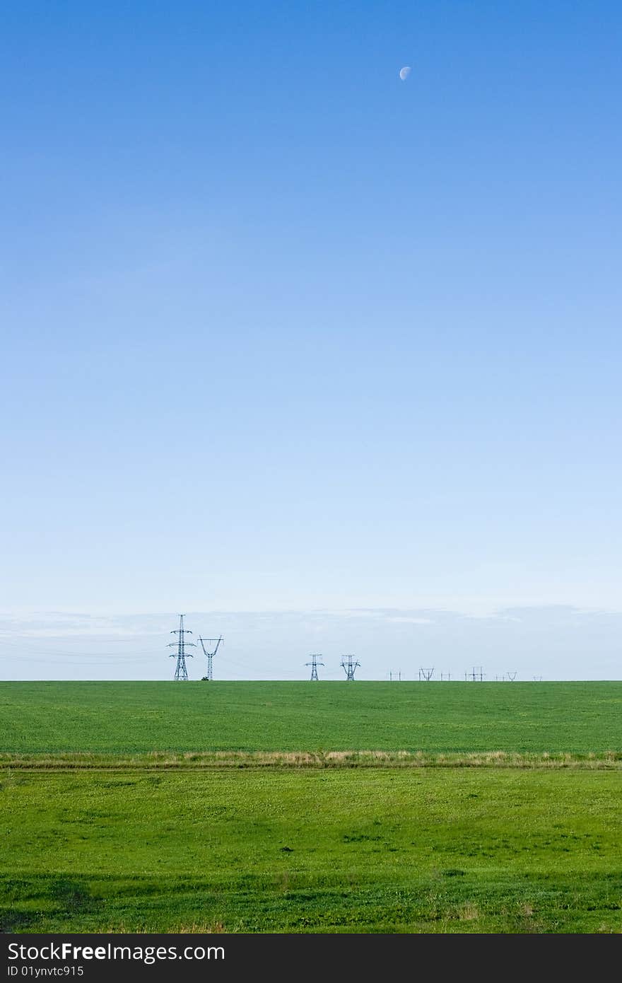 Green field and blue sky. Moon in the sky. Power lines are visible on the horizon.