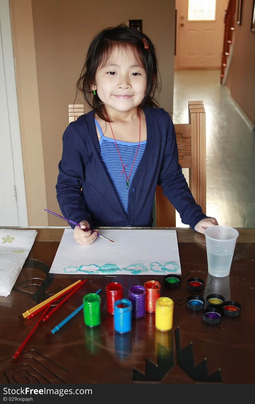 Asian girl painting at a table with paints in front of her and a paint brush inside her hand. Asian girl painting at a table with paints in front of her and a paint brush inside her hand