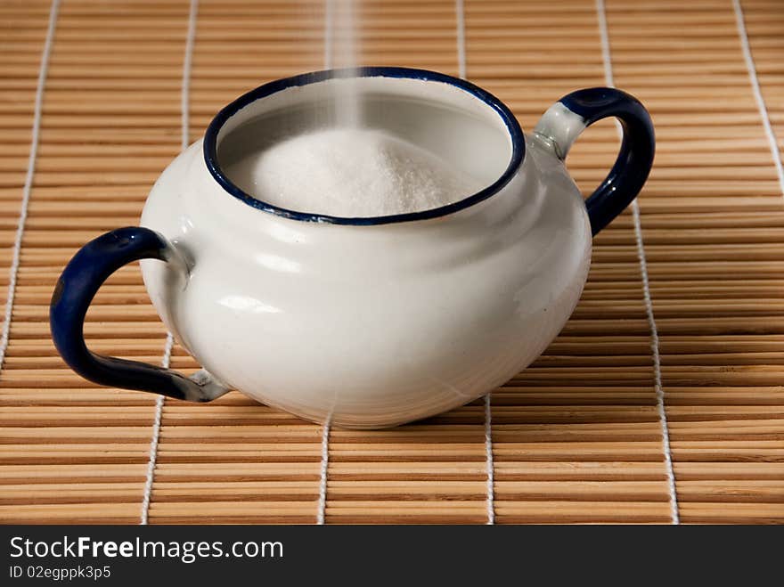 A white enamel sugar bowl on a straw mat being filled with sugar. A white enamel sugar bowl on a straw mat being filled with sugar