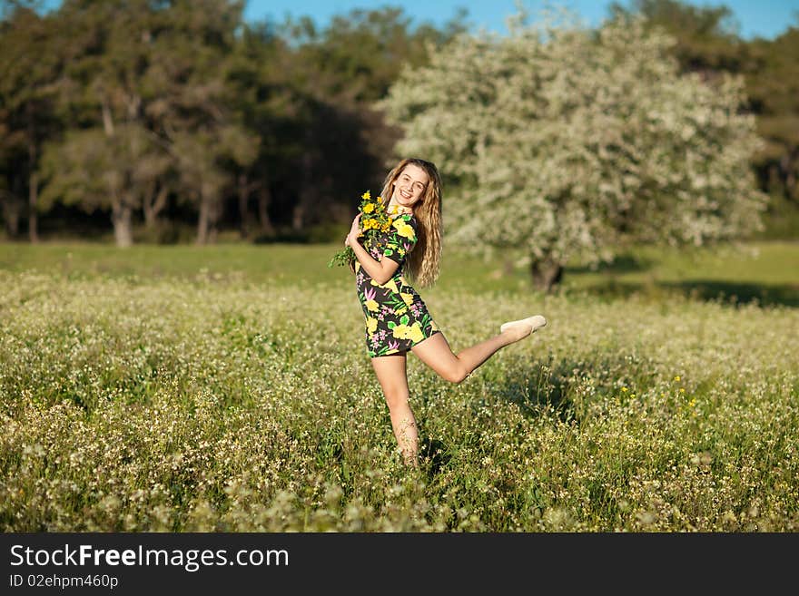 Beautiful young blonde woman jumping in blooming meadow in spring, bunch of yellow flowers in hand; shallow depth of field, trees in background. Beautiful young blonde woman jumping in blooming meadow in spring, bunch of yellow flowers in hand; shallow depth of field, trees in background