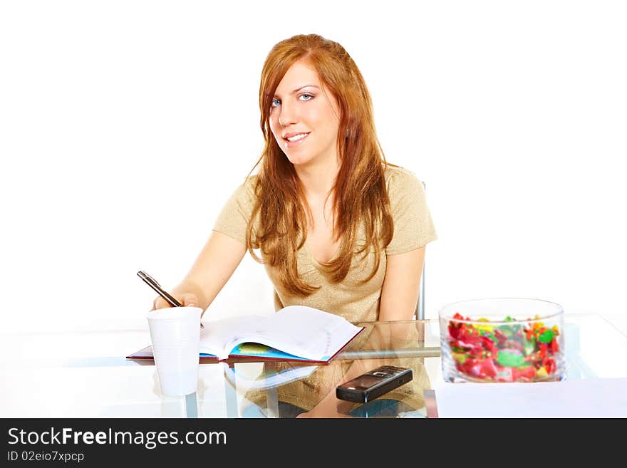 Smiling student girl learning at desk with notebook with pen in hand, mobile phone and some sweets. Smiling student girl learning at desk with notebook with pen in hand, mobile phone and some sweets