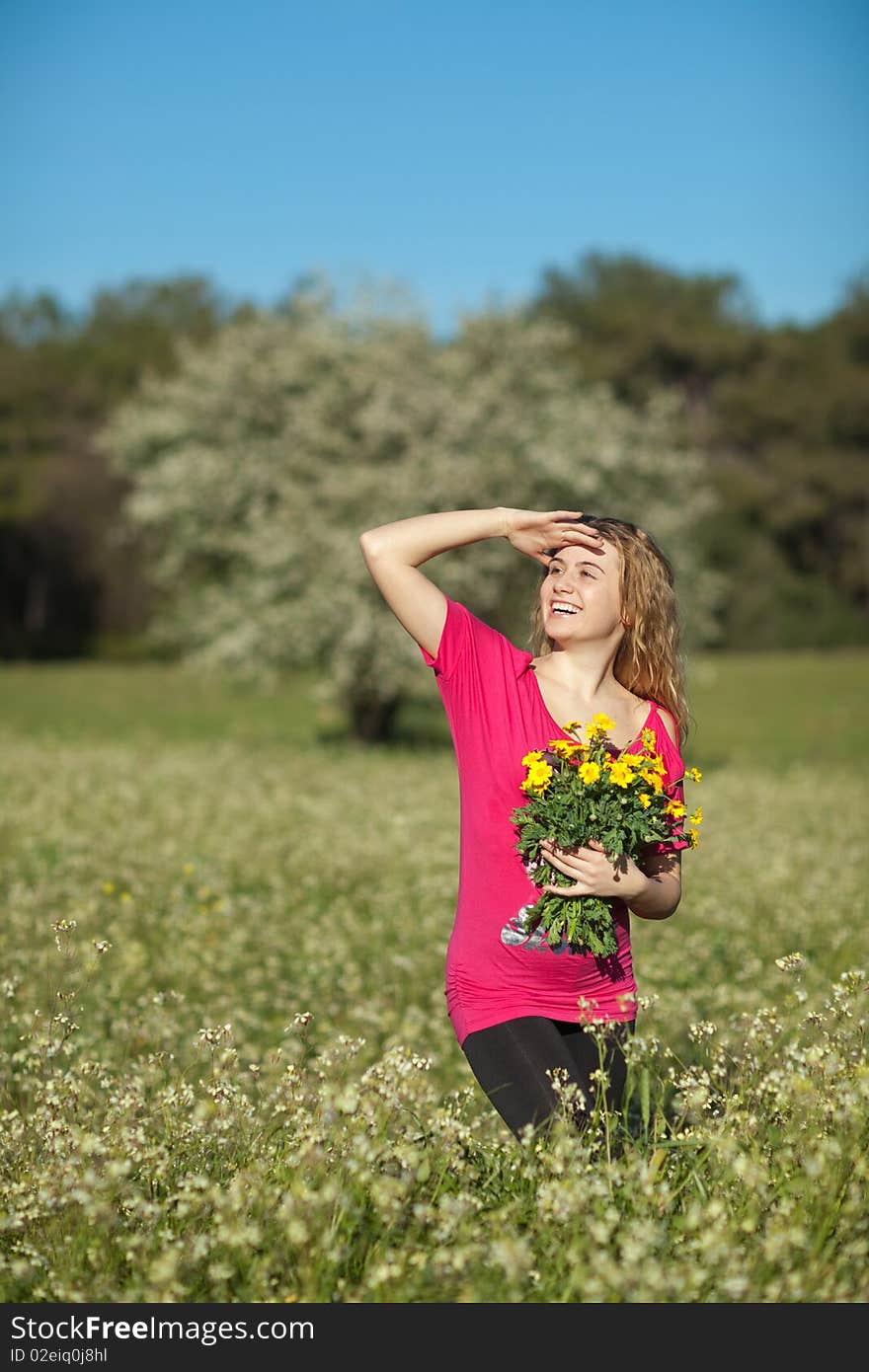 Beautiful young blonde woman standing in blooming meadow in spring, bunch of yellow flowers in hand, smiling; shallow depth of field, trees in background. Beautiful young blonde woman standing in blooming meadow in spring, bunch of yellow flowers in hand, smiling; shallow depth of field, trees in background