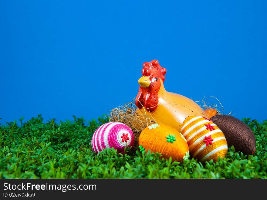 Handmade easter egg and ceramic hen placed on cress. Small eggs are covered by cotton string in different colours. Additional decorative elements are glued to egg. Blue background. Ceramic hen is used as Easter decoration. Handmade easter egg and ceramic hen placed on cress. Small eggs are covered by cotton string in different colours. Additional decorative elements are glued to egg. Blue background. Ceramic hen is used as Easter decoration