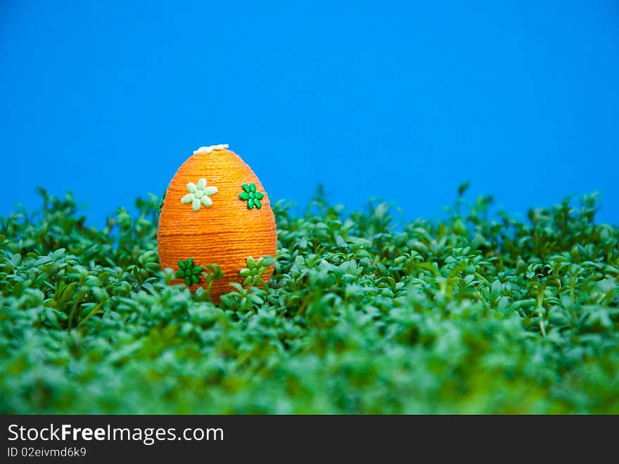 Handmade easter egg dropped on cress. Small eggs are covered by cotton string in different colours. Additional decorative elements are glued to egg. Blue background. Handmade easter egg dropped on cress. Small eggs are covered by cotton string in different colours. Additional decorative elements are glued to egg. Blue background.