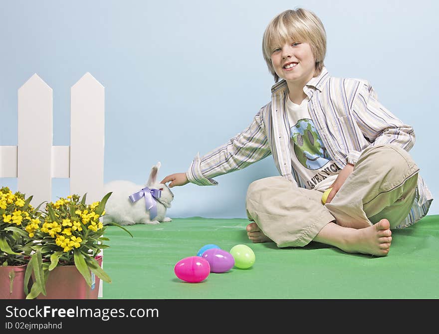 Blond, seated boy reaches out to pet a white rabbit wearing a bow. Blond, seated boy reaches out to pet a white rabbit wearing a bow