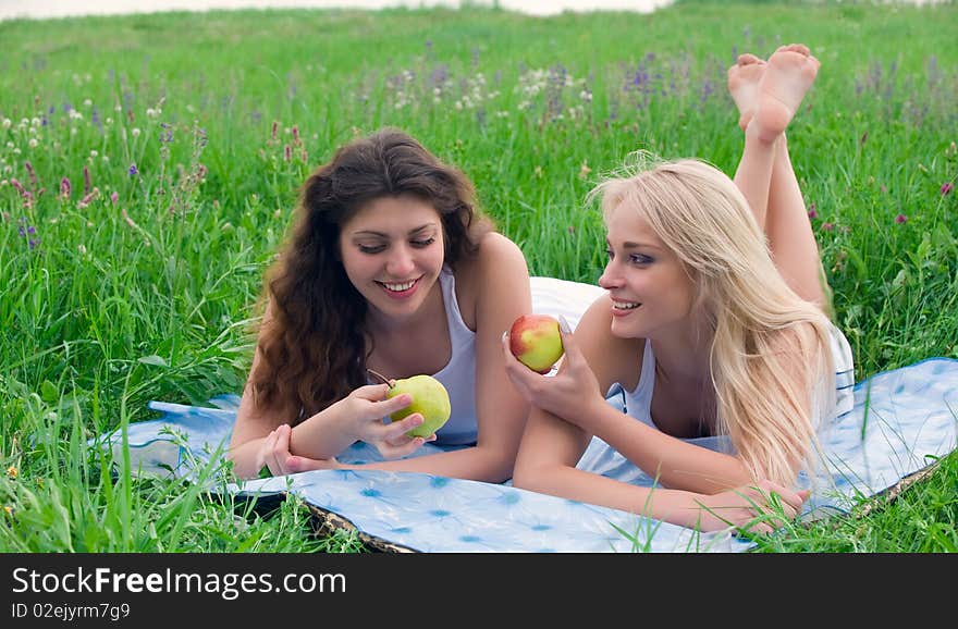 Two attractive girls blonde and brunette with long hair lying on the rug on the grass with an apple in the hands. Two attractive girls blonde and brunette with long hair lying on the rug on the grass with an apple in the hands