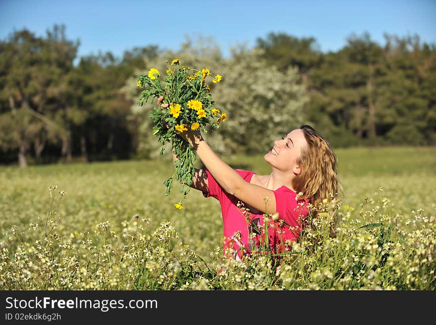 Beautiful young blonde woman standing in blooming meadow in spring, bunch of yellow flowers in hand, smiling, blue sky and trees in background; shallow depth of field. Beautiful young blonde woman standing in blooming meadow in spring, bunch of yellow flowers in hand, smiling, blue sky and trees in background; shallow depth of field