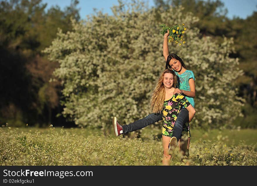 Two beautiful young women standing in blooming meadow in spring, giving piggyback, bunch of yellow flowers in hand, smiling, tree in background; shallow depth of field. Two beautiful young women standing in blooming meadow in spring, giving piggyback, bunch of yellow flowers in hand, smiling, tree in background; shallow depth of field