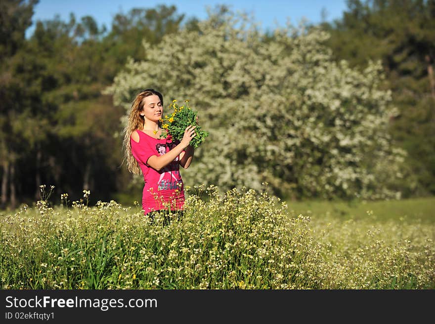 Beautiful young blonde woman standing in blooming meadow in spring, bunch of yellow flowers in hand, smelling eyes closed, blue sky and trees in background; shallow depth of field. Beautiful young blonde woman standing in blooming meadow in spring, bunch of yellow flowers in hand, smelling eyes closed, blue sky and trees in background; shallow depth of field