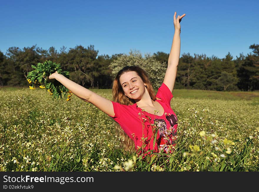 Beautiful young blonde woman standing in blooming meadow in spring, bunch of yellow flowers in hand, cheerful, smiling, blue sky and trees in background; shallow depth of field. Beautiful young blonde woman standing in blooming meadow in spring, bunch of yellow flowers in hand, cheerful, smiling, blue sky and trees in background; shallow depth of field