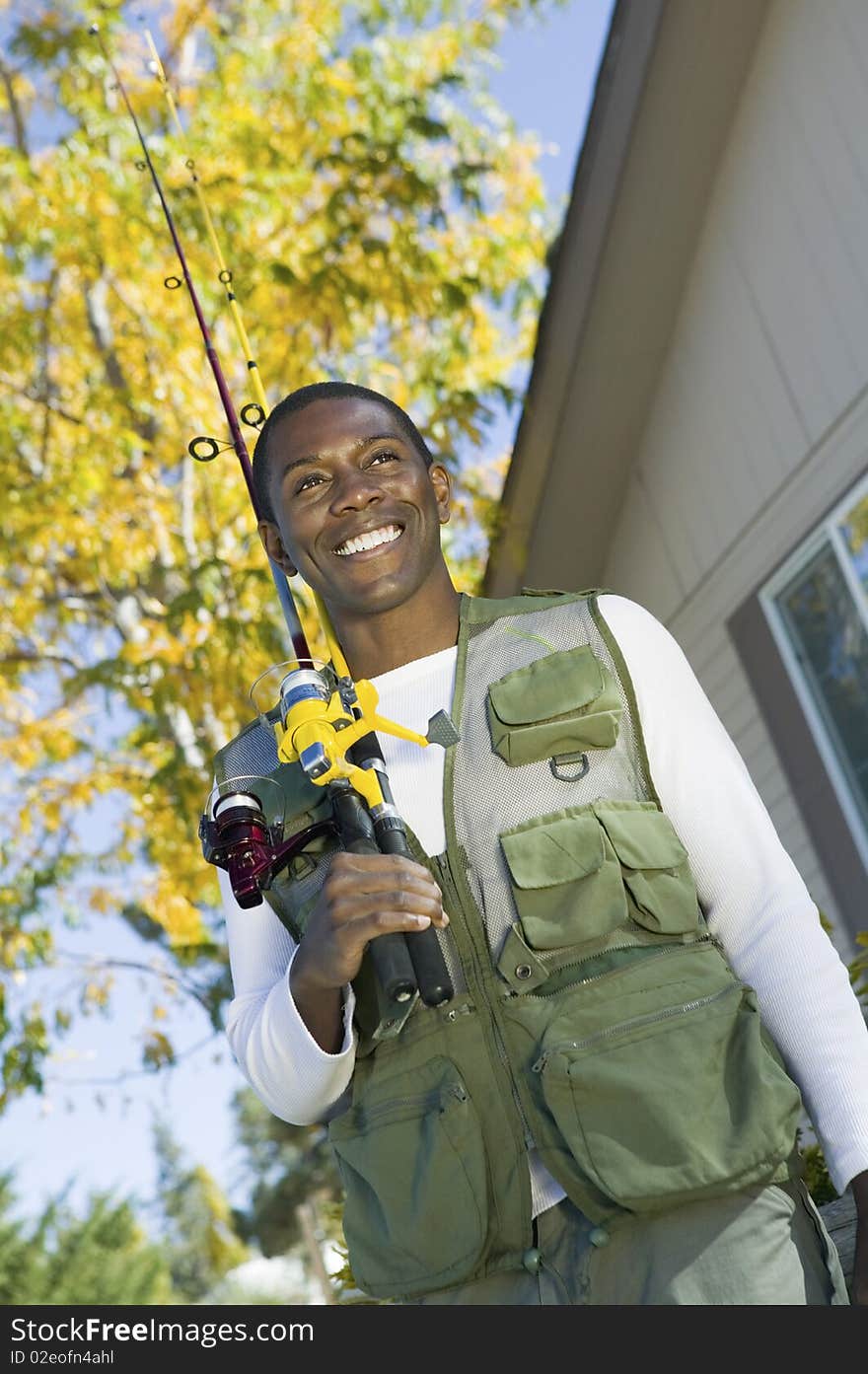 Man holding fishing rod in font of house, smiling, (low angle view)