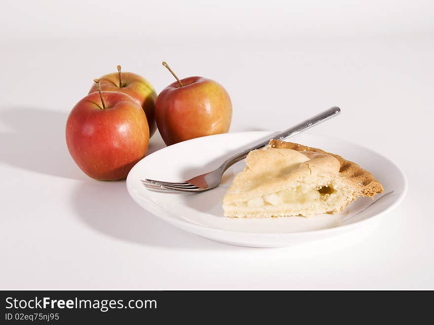 A horizontal image of a slice of apple pie on a white plate with three red apples in the background