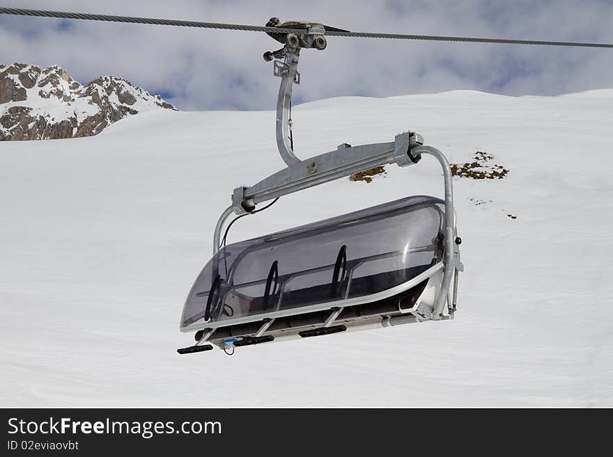 Empty ski chair lift on a cable with a snowy rocky mountain in the background against a cloudy blue sky. Empty ski chair lift on a cable with a snowy rocky mountain in the background against a cloudy blue sky