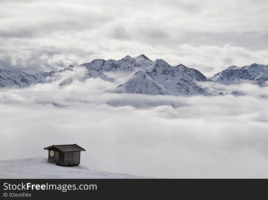 Wooden cabin above the clouds overlooking snowy mountain peaks with a cloudy sky